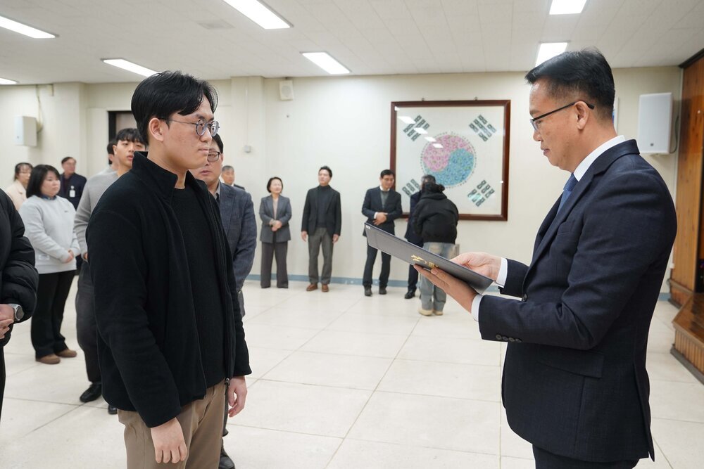 2026년 상반기 4급이하 정기인사로 정장을 입고 안경을 쓴 남성이 실내 행사장에서 한 남성에게 상장을 전달하며 내용을 읽고 있고, 남성은 검은 자켓을 입고 서 있으며 뒤쪽에는 참석자들이 줄지어 서서 행사를 지켜보고 있다.
