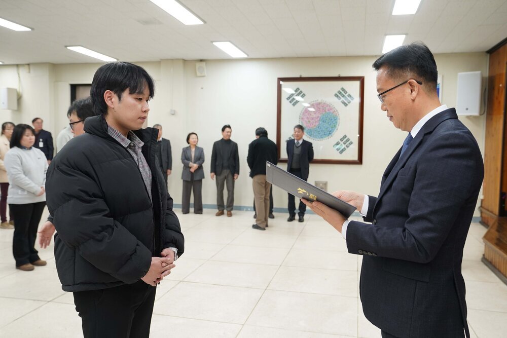 2026년 상반기 4급이하 정기인사로 정장을 입고 안경을 쓴 남성이 실내 행사장에서 한 남성에게 상장을 전달하며 내용을 읽고 있고, 남성은 검은 패딩을 입고 두 손을 모은 채 서 있으며 뒤쪽에는 참석자들이 줄지어 서서 행사를 지켜보고 있다.
