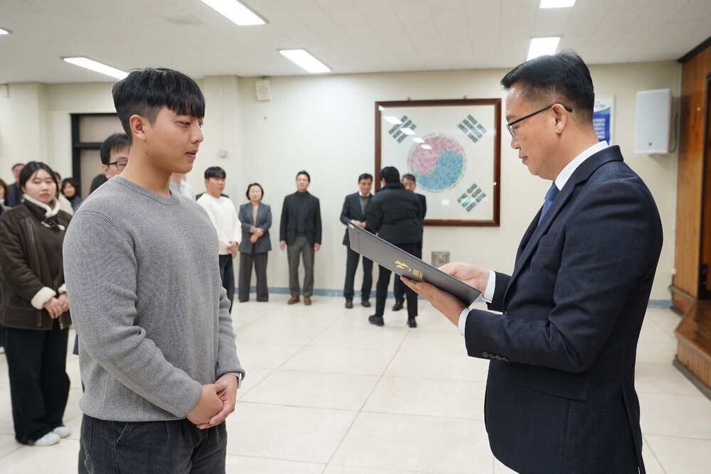 2026년 상반기 4급이하 정기인사로 정장을 입고 안경을 쓴 남성이 실내 행사장에서 한 남성에게 상장을 전달하며 내용을 읽고 있고, 남성은 회색 니트를 입고 두 손을 모은 채 서 있으며 뒤쪽에는 참석자들이 줄지어 서서 행사를 지켜보고 있다.