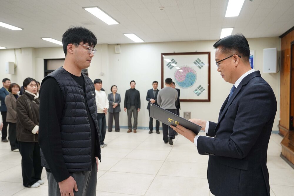 2026년 상반기 4급이하 정기인사로 정장을 입고 안경을 쓴 남성이 실내 행사장에서 한 남성에게 상장을 전달하며 내용을 읽고 있고, 남성은 검은 패딩 조끼를 입고 서 있으며 뒤쪽에는 참석자들이 줄지어 서서 행사를 지켜보고 있다.