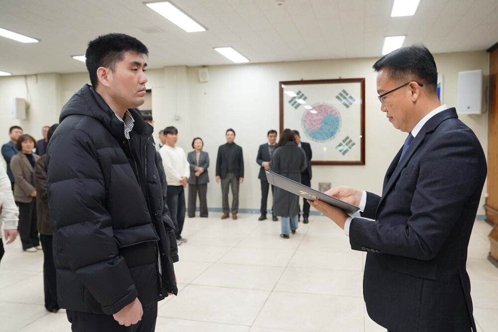 2026년 상반기 4급이하 정기인사로 정장을 입고 안경을 쓴 남성이 실내 행사장에서 한 남성에게 상장을 전달하며 내용을 읽고 있고, 남성은 검은 패딩을 입고 서 있으며 뒤쪽에는 참석자들이 줄지어 서서 행사를 지켜보고 있다.