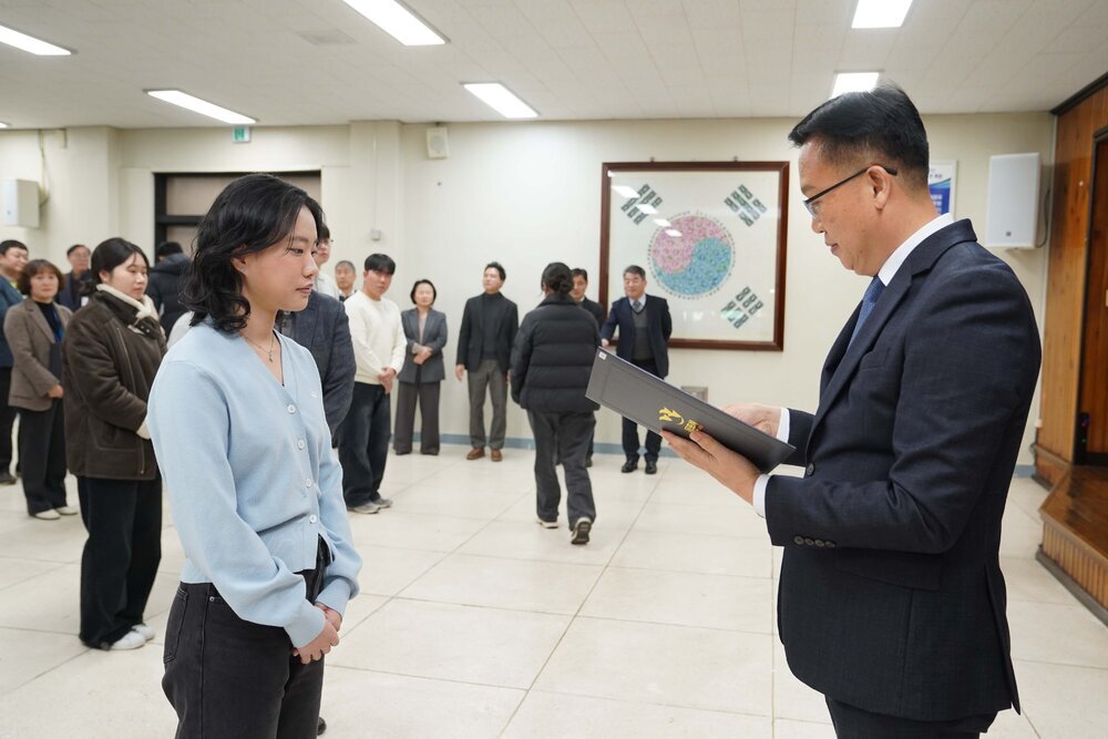 2026년 상반기 4급이하 정기인사로 정장을 입고 안경을 쓴 남성이 실내 행사장에서 한 여성에게 상장을 전달하며 내용을 읽고 있고, 여성은 하늘색 가디건을 입고 두 손을 모은 채 서 있으며 뒤쪽에는 참석자들이 줄지어 서서 행사를 지켜보고 있다.