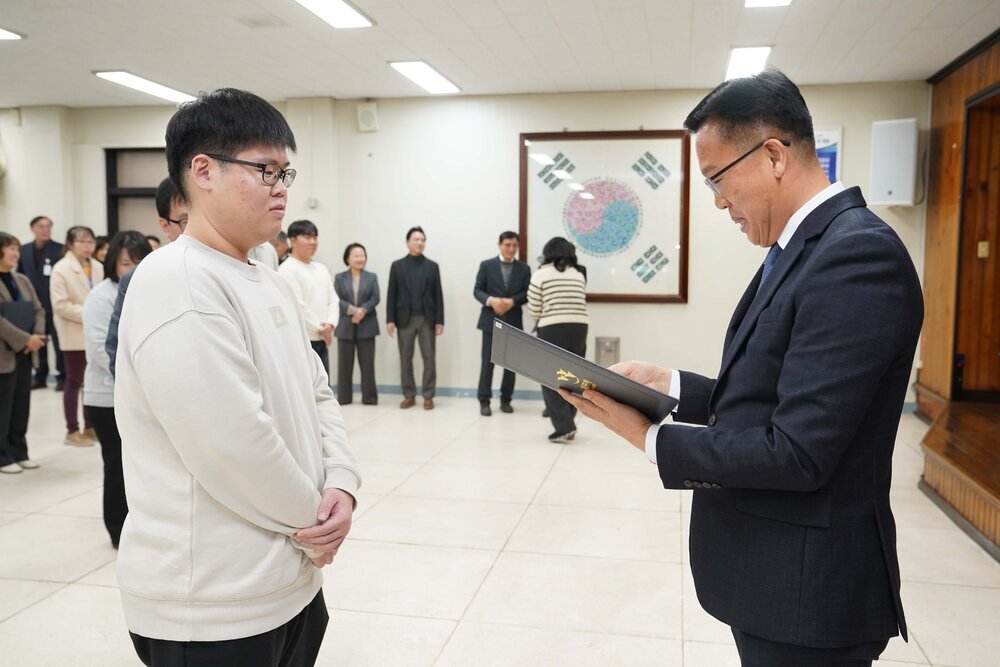 2026년 상반기 4급이하 정기인사로 정장을 입고 안경을 쓴 남성이 실내 행사장에서 한 남성에게 상장을 전달하며 내용을 읽고 있고, 남성은 밝은색 맨투맨을 입고 두 손을 모은 채 서 있으며 뒤쪽에는 참석자들이 줄지어 서서 행사를 지켜보고 있다.