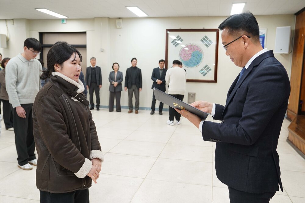 2026년 상반기 4급이하 정기인사로 정장을 입고 안경을 쓴 남성이 실내 행사장에서 한 여성에게 상장을 전달하며 내용을 읽고 있고, 여성은 갈색 무스탕 코트를 입고 두 손을 모은 채 앞에 서 있으며 뒤쪽에는 여러 사람들이 줄지어 서서 행사를 지켜보고 있다.