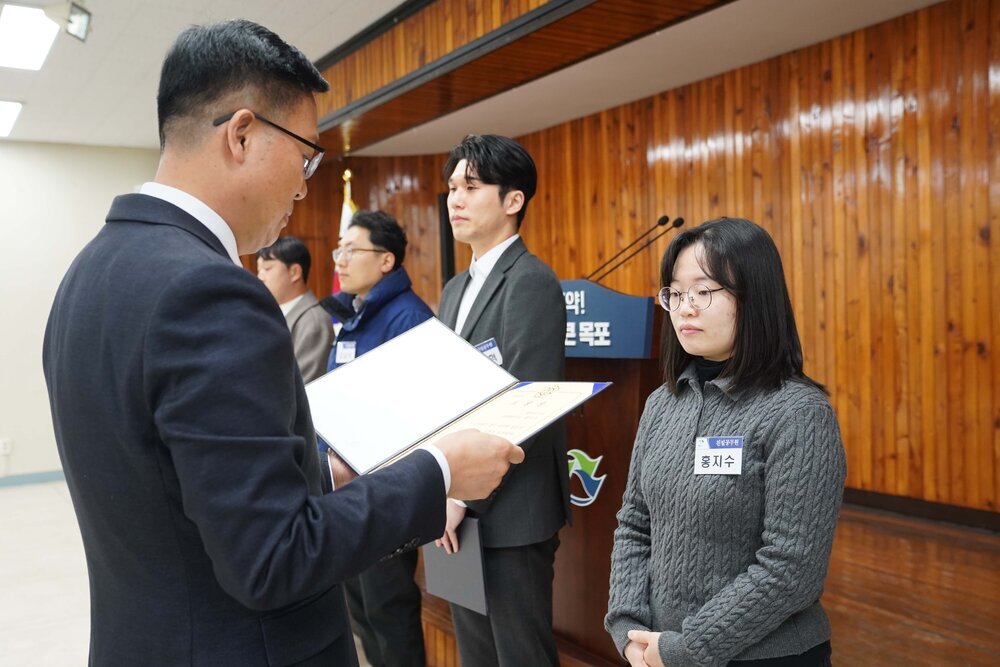 2월 정례조회에 정장을 입고 안경을 낀 남성이 어꺠보다 약간 긴 단발머리에 검은색 목티에 짙은 회색 꽈베기니트를 입은 홍지수에게 패를 수여하고 있다.