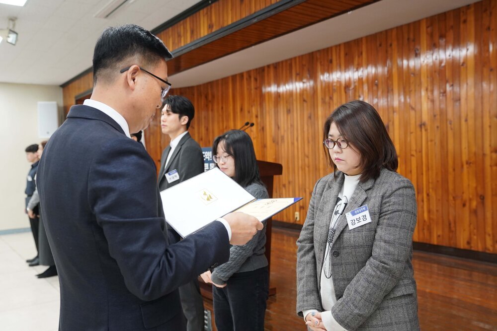 2월 정례조회에 정장을 입고 안경을 낀 남성이 목까지오는 단발머리에 흰색 티에 회색 체크무늬 자켓을 입은 강보경에게 패를 수여하고 있다.