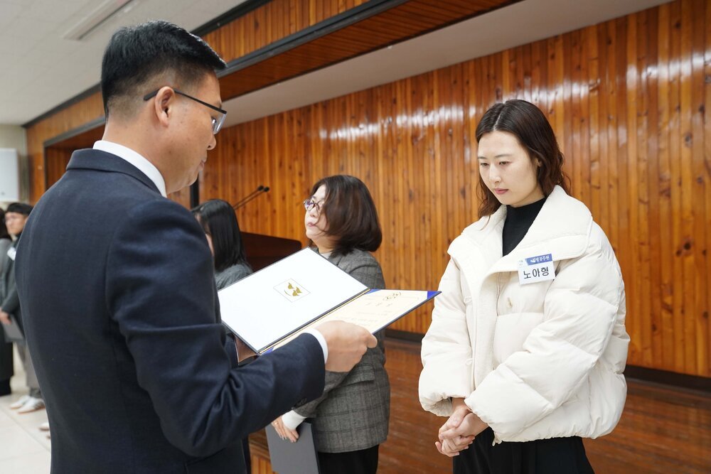 2월 정례조회에 정장을 입고 안경을 낀 남성이 긴 파마머리에 밝은 베이지 숏패딩을 입은 노아형에게 패를 수여하고 있다.
