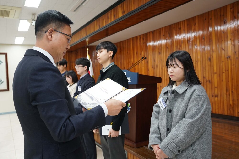 2월 정례조회에 정장을 입고 안경을 낀 남성이 밝은 회색 코트를 정은애에게 패를 수여하고 있다.