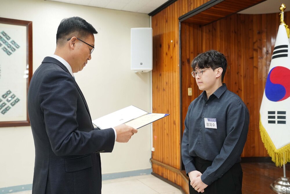 2월 정례조회에 정장을 입고 안경을 낀 남성이 검은 바지에 짙은 네이비 셔츠를 입은 정지수에게 패를 수여하고 있다.