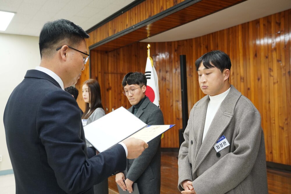 2월 정례조회에 정장을 입고 안경을 낀 남성이 흰색 목티에 밝은 오트밀색 코트를 입은 최성관에게 패를 수여하고 있다.