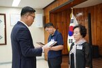 정장 차림의 관계자가 검은색 상의와 스카프를 착용한 여성 수상자에게 패를 전달하며 읽고 있는 모습이다.