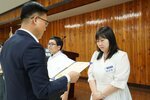 정장 차림의 관계자가 흰색 원피스를 입은 여성 수상자에게 패를 전달하며 읽고 있는 모습이다.