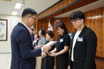 정장 차림의 관계자가 검은색 재킷을 입은 남성 수상자에게 패를 전달하며 읽고 있는 모습이다.