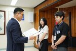 정장 차림의 관계자가 검은색 반팔 상의를 입은 남성 수상자에게 패를 전달하며 읽고 있는 모습이다.