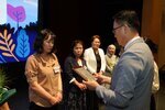 정장 차림의 관계자가 베이지색 블라우스를 입은 여성 수상자에게 패를 전달하며 읽고 있는 모습이다.
