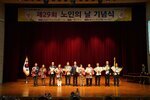 노인의 날 기념식에서 수상자들이 꽃다발을 들고 무대 위에서 단체 기념 촬영을 하고 있는 모습이다.