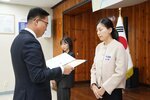 11월 정례조회로 정장을 입고 안경을 쓴 남성이 긴머리를 뒤로 묵고 밝은 베이지색 짧은 코트를 입은 강미혜 수상자에게 상장을 수여 하고 있다.