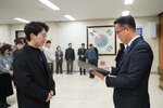 2026년 상반기 4급이하 정기인사로 정장을 입은 남성이 실내 행사장에서 한 남성에게 상장을 전달하며 내용을 읽고 있고, 남성은 안경을 쓰고 검은색 코트를 입고 사원증을 착용한 채 두 손을 모은 상태로 서 있으며 뒤쪽으로 참석자들이 줄지어 서서 행사를 지켜보고 있다.