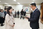 2026년 상반기 4급이하 정기인사로 정장을 입은 남성이 실내 행사장에서 한 여성에게 상장을 전달하며 내용을 읽고 있고, 여성은 밝은 베이지색 코트를 입고 사원증을 착용한 채 두 손을 모은 상태로 서 있으며 뒤쪽으로 참석자들이 줄지어 서서 행사를 지켜보고 있다.