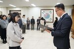 2026년 상반기 4급이하 정기인사로 정장을 입은 남성이 실내 행사장에서 한 여성에게 상장을 전달하며 내용을 읽고 있고, 여성은 안경을 쓰고 밝은 회색 카라 자켓을 입고 사원증을 착용한 채 두 손을 모은 상태로 서 있으며 뒤쪽으로 참석자들이 줄지어 서서 행사를 지켜보고 있다.