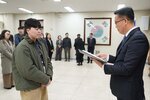 2026년 상반기 4급이하 정기인사로 정장을 입은 남성이 실내 행사장에서 한 남성에게 상장을 전달하며 내용을 읽고 있고, 남성은 카키색 짧은 패딩을 입고 두 손을 모은 상태로 서 있으며 뒤쪽으로 참석자들이 줄지어 서서 행사를 지켜보고 있다.