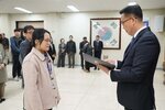 2026년 상반기 4급이하 정기인사로 정장을 입은 남성이 실내 행사장에서 한 여성에게 상장을 전달하며 내용을 읽고 있고, 여성은 밝은 베이지색 스웨이드 자켓을 입고 사원증을 착용한 채 두 손을 내린 상태로 서 있으며 뒤쪽에는 참석자들이 줄지어 서서 행사를 지켜보고 있다.