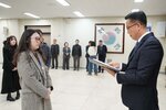 2026년 상반기 4급이하 정기인사로 정장을 입은 남성이 실내 행사장에서 한 여성에게 상장을 전달하며 내용을 읽고 있고, 여성은 긴 파마머리에 밝은 회색과 베이지가 썩힌 체크 코드를 입고 사원증을 착용한 채 두 손을 모은 상태로 서 있으며 뒤쪽에는 참석자들이 줄지어 서서 행사를 지켜보고 있다.