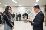 2026년 상반기 4급이하 정기인사로 정장을 입은 남성이 실내 행사장에서 한 여성에게 상장을 전달하며 내용을 읽고 있고, 여성은 긴 생머리에 검은생 티에 회색 목까지올라오는 베스트를 입고 사원증을 착용한 채 두 손을 모은 상태로 서 있으며 뒤쪽에는 참석자들이 줄지어 서서 행사를 지켜보고 있다.