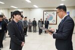2026년 상반기 4급이하 정기인사로 정장을 입은 남성이 실내 행사장에서 한 남성에게 상장을 전달하며 내용을 읽고 있고, 남성은 검은 정장을 입은 채 두 손을 모은 상태로 서 있으며 뒤쪽에는 참석자들이 줄지어 서서 행사를 지켜보고 있다.