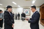 2026년 상반기 4급이하 정기인사로 정장을 입은 남성이 실내 행사장에서 한 남성에게 상장을 전달하며 내용을 읽고 있고, 남성은 검은 패딩 점퍼를 입은 채 두 손을 모은 상태로 서 있으며 뒤쪽에는 참석자들이 줄지어 서서 행사를 지켜보고 있다.