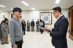 2026년 상반기 4급이하 정기인사로 정장을 입은 남성이 실내 행사장에서 한 남성에게 상장을 전달하며 내용을 읽고 있고, 남성은 회색 니트를 입은 채 두 손을 모은 상태로 서 있으며 뒤쪽에는 참석자들이 줄지어 서서 행사를 지켜보고 있다.