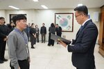 2026년 상반기 4급이하 정기인사로 정장을 입은 남성이 실내 행사장에서 한 남성에게 상장을 전달하며 내용을 읽고 있고, 남성은 회색 셔츠와 사원증을 착용한 채 서 있으며 뒤쪽에는 참석자들이 줄지어 서서 행사를 지켜보고 있다.