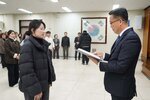 2026년 상반기 4급이하 정기인사로 정장을 입고 안경을 쓴 남성이 실내 행사장에서 한 여성에게 상장을 전달하며 내용을 읽고 있고, 여성은 검은 패딩을 입고 두 손을 모은 채 서 있으며 뒤쪽에는 참석자들이 줄지어 서서 행사를 지켜보고 있다.