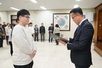 2026년 상반기 4급이하 정기인사로 정장을 입고 안경을 쓴 남성이 실내 행사장에서 한 남성에게 상장을 전달하며 내용을 읽고 있고, 남성은 밝은색 맨투맨을 입고 두 손을 모은 채 서 있으며 뒤쪽에는 참석자들이 줄지어 서서 행사를 지켜보고 있다.