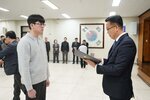 2026년 상반기 4급이하 정기인사로 정장을 입고 안경을 쓴 남성이 실내 행사장에서 한 남성에게 상장을 전달하며 내용을 읽고 있고, 남성은 회색 니트를 입고 정면을 바라보며 서 있으며 뒤쪽에는 여러 사람들이 줄지어 서서 행사를 지켜보고 있다.