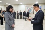 2026년 상반기 4급이하 정기인사로 정장을 입고 안경을 쓴 남성이 실내 행사장에서 한 여성에게 상장을 전달하며 내용을 읽고 있고, 여성은 회색 코트를 입고 두 손을 모은 채 서 있으며 뒤쪽에는 여러 사람들이 줄지어 서서 행사를 지켜보고 있다.