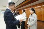 2월 정례조회에 정장을 입고 안경을 낀 남성이 긴 파마머리를 뒤로 묶고 밝은 회색 긴 코트를 입은 현승아에게 패를 수여하고 있다.